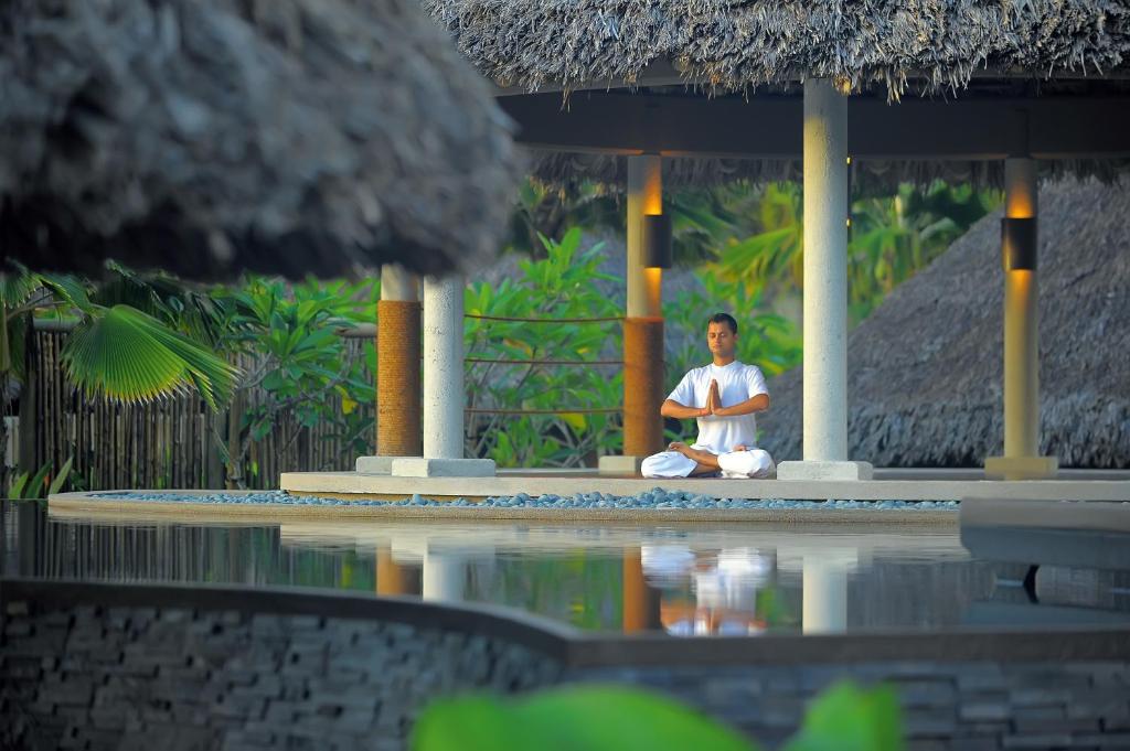 Meditējošs cilvēks mierīgā vidē, apņemts ar zaļumu un ūdens atspulgu, veicina relaksāciju un harmoniju.