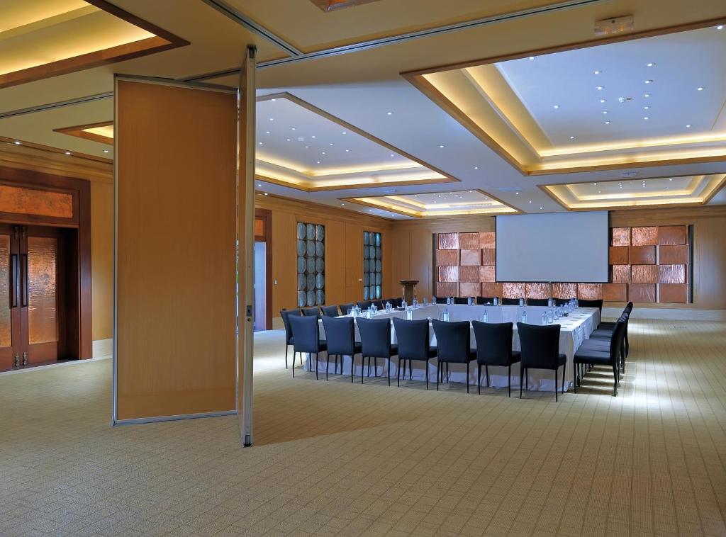 Konferenču telpa ar apaļo galdu, melnām krēsliem un lielu ekrānu prezentācijām. Telpa ir gaiša un moderna, ar elegantu apgaismojumu un labiekārtojumu.