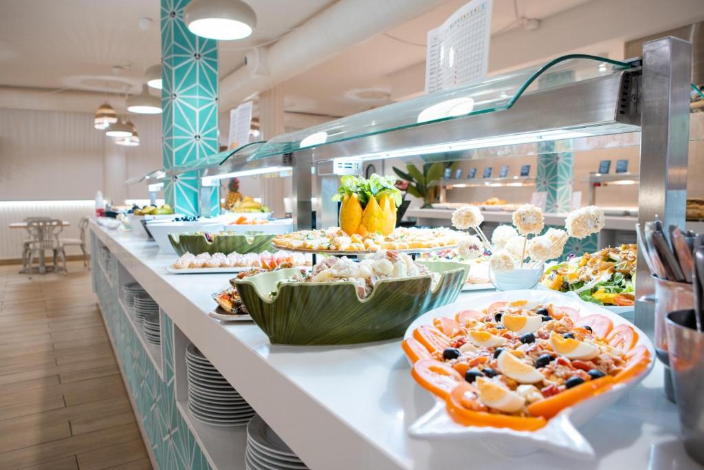 Bagātīgs bufetes galds ar dažādiem ēdieniem, tostarp salātiem, desertiem un augļiem, izvietots gaišā un mūsdienīgā telpā. Ideal for a vibrant dining experience.