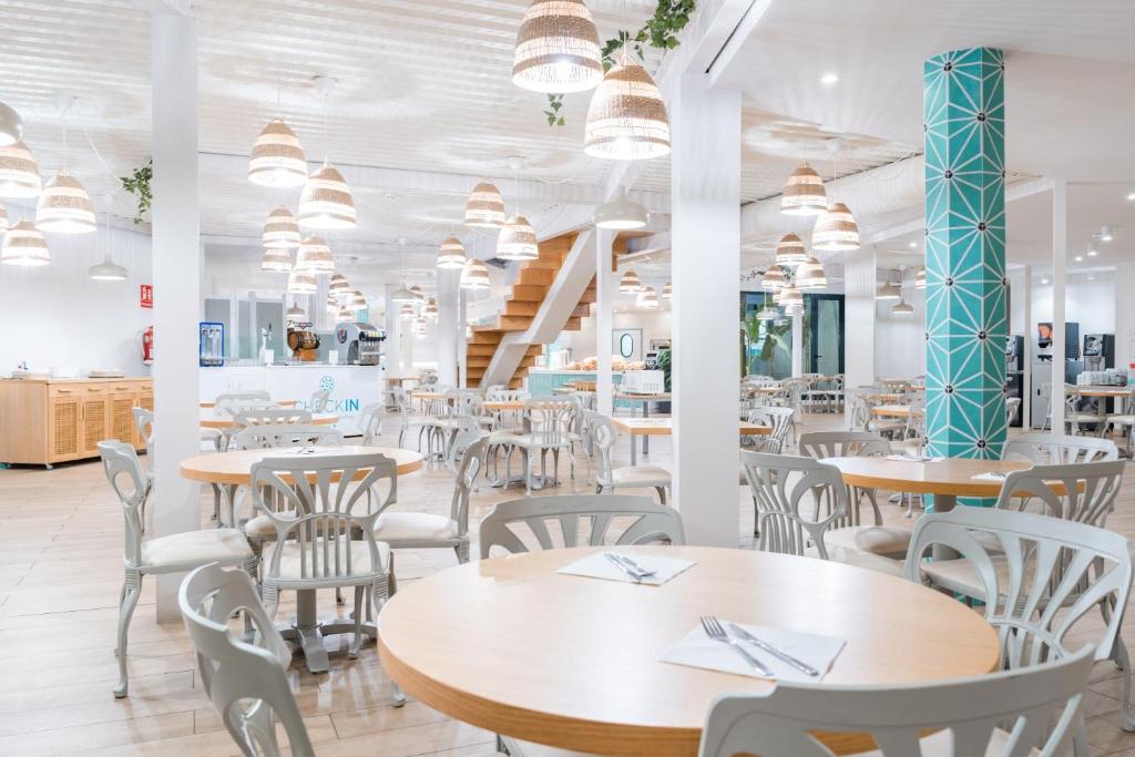 Moderns restorāns ar gaišu interjeru, koka galdiem un ērtām krēslām, apgaismots ar stilīgām piekaramajām lampām. Telpā ir redzami augi un dekoratīvi elementi, radot patīkamu atmosfēru.