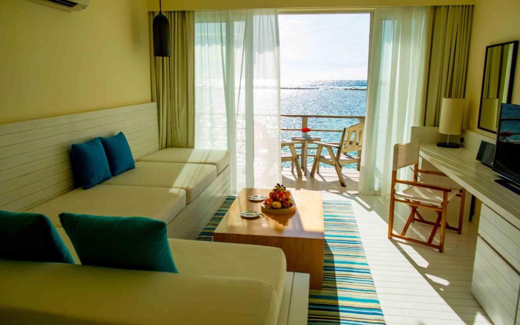 Mūsdienīgs viesnīcas numurs ar skatu uz jūru, kurā ir ērti dīvāni, koka galds ar augļiem un balkons. Gaisma ieplūst caur logiem, radot siltu un relaksējošu atmosfēru.