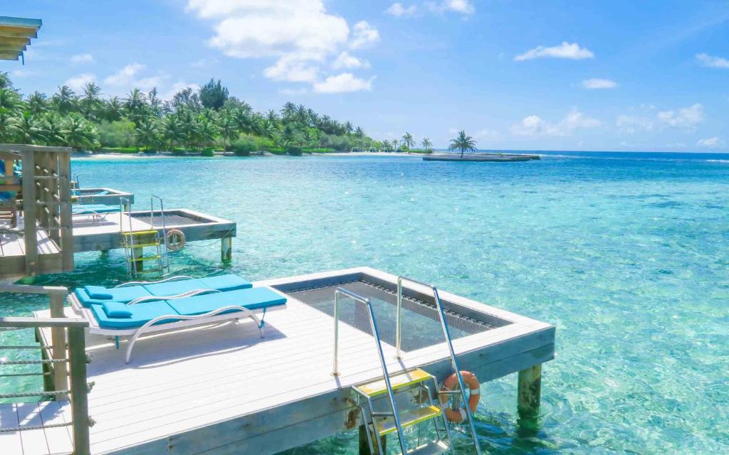 Skaista pludmale ar skaidru ūdeni un palmām, kas sniedz idillisku atpūtas vidi. Uz koka klāja atrodas sauļošanās krēsli, kas aicina uz relaksāciju.