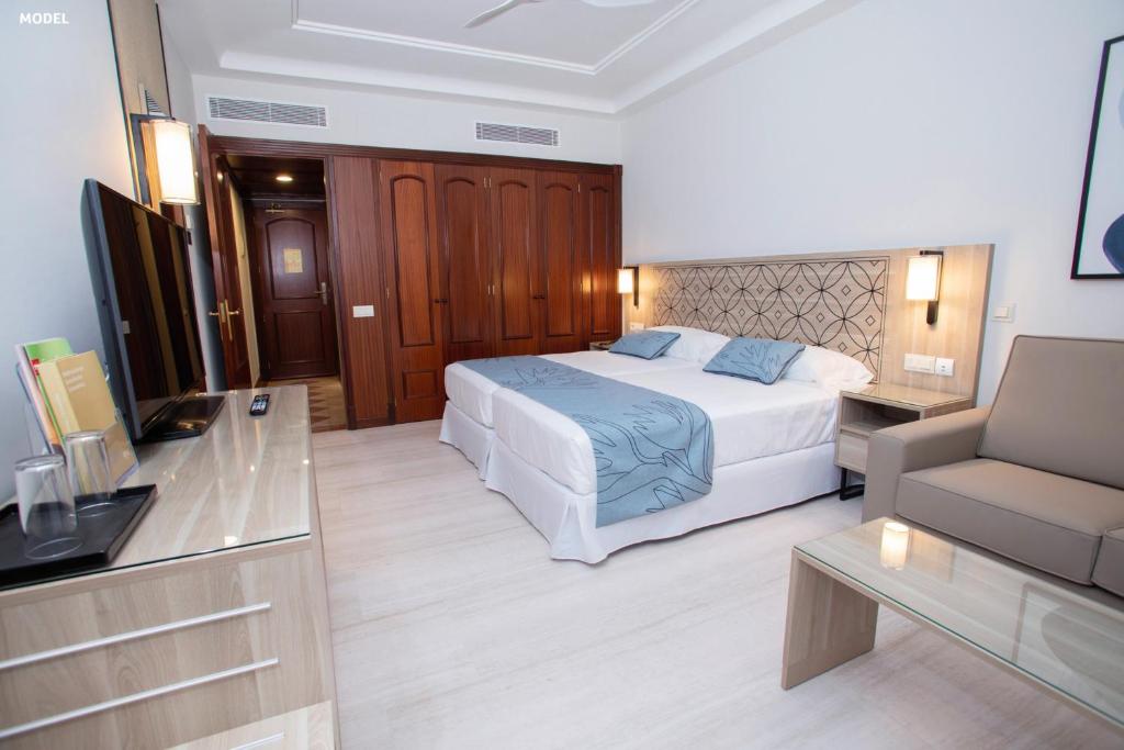 Mūsdienīga viesnīcas istaba ar divguļamo gultu, ērtu dīvānu un televizoru, nodrošinot komfortablus apstākļus atpūtai. Gaisma un neitrālas krāsas rada patīkamu atmosfēru.