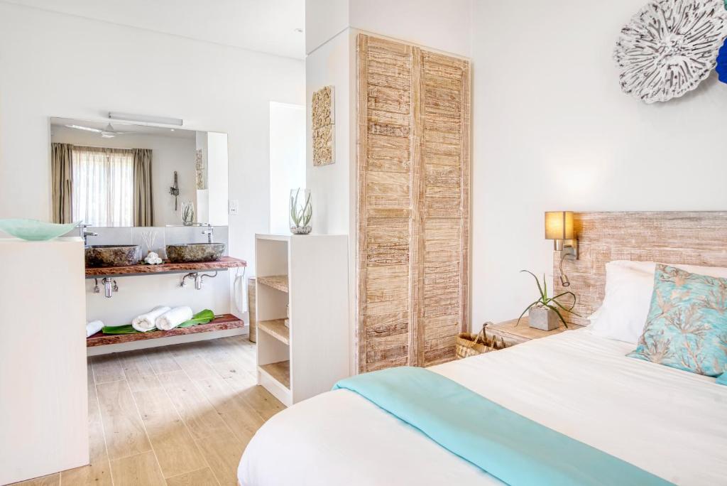 Mūsdienīgs guļamistabas interjers ar atvērtu vannas istabu, kas ietver divas akmens izlietnes un dabīgu apgaismojumu. Svaigi balti paklāji un vienkāršs, elegants dizains rada relaksējošu atmosfēru.