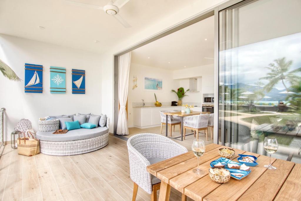 Mūsdienīgs viesistabas interjers ar atvērtiem logiem, ērtu sēdvietu un ēdamgaldu, kas piedāvā skatu uz dabu. Dekorēts ar jūras tematiku un svaigām krāsām, radot relaksējošu atmosfēru.