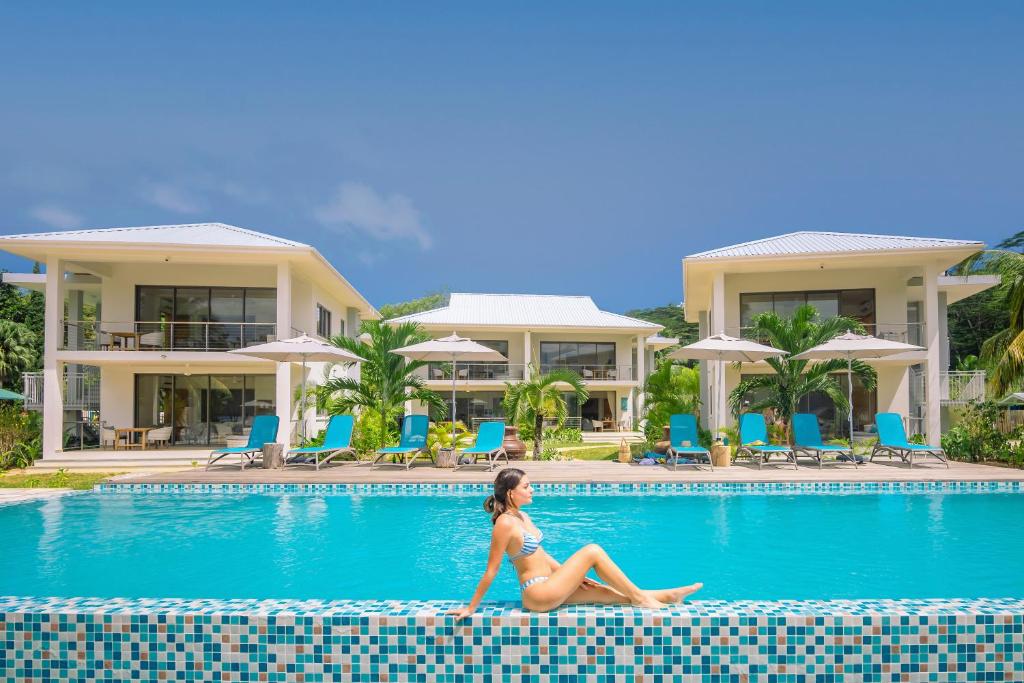 Peldbaseins ar modernu arhitektūru un atpūtas zonām, kur sieviete relaksējas pie ūdens. Šī ainava rada mierīgu un atpūtu veicinošu atmosfēru.