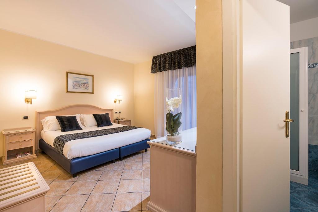 Mūsdienīga viesnīcas istaba ar divguļamo gultu, elegantu apgaismojumu un logu ar aizkariem. Istabā ir koka mēbeles un ziedu kompozīcija uz galda.