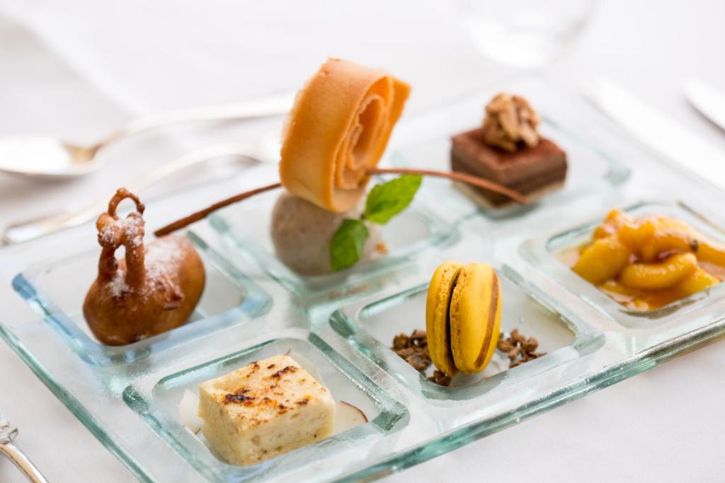 Deserts uz stikla šķīvja, kas satur dažādas delikateses, tostarp šokolādes kvadrātiņu, makaronu, augļu mērci un riekstu dekorāciju.