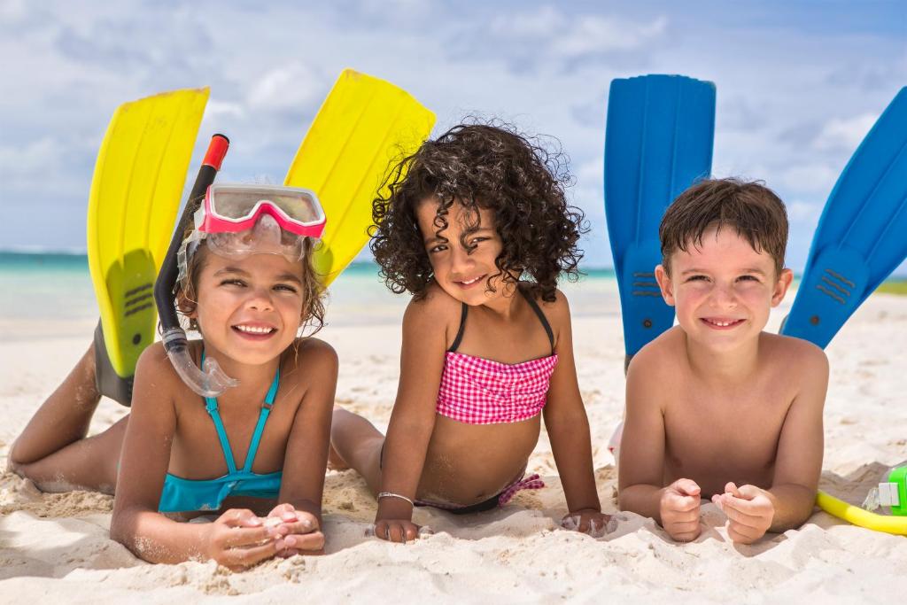 Trīs bērni atpūšas pludmalē, ģērbušies peldkostīmos un ar snorkelēšanas aprīkojumu, smaidot uz smilšainā fona. Aiz viņiem redzami peldēšanas zābaki.