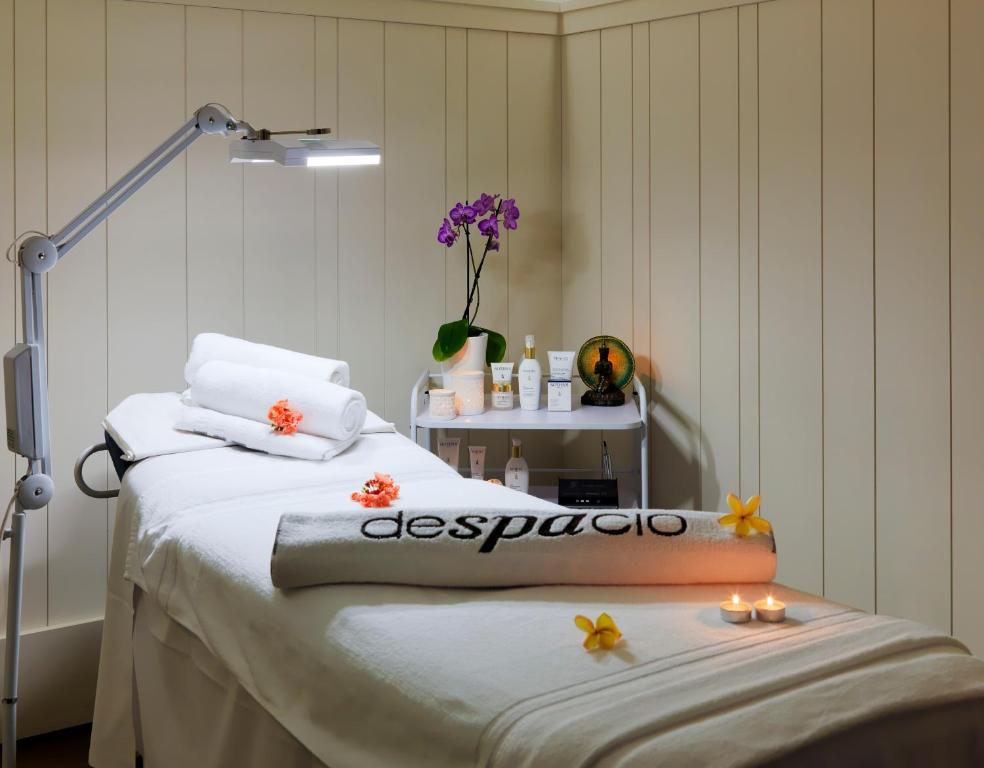 Mierīgā spa telpa ar masāžas galdu, uz kura novietoti dvieļi un ziedi. Telpā ir relaksējošs apgaismojums un aromātiskas svecītes, radot nomierinošu atmosfēru.