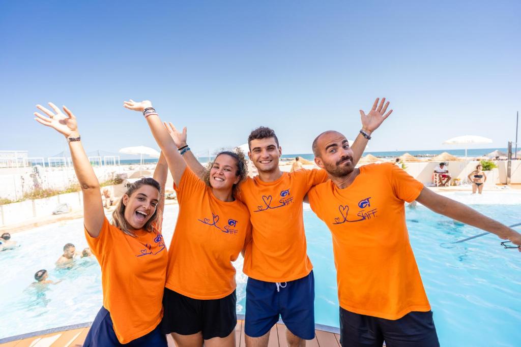 Priecīgs grupas foto ar cilvēkiem oranžās t-kreklos, kas pozē pie baseina, baudot saulainu dienu. Fons ietver peldētājus un pludmales sauļošanās zonas.