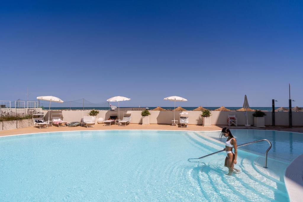 Peldbaseins ar skatu uz jūru, kur sieviete tīra ūdeni, apņemta ar sauļošanās krēsliem un saules paraplājiem. Izsmalcināta atpūtas vieta ar skaidru debesi un mierīgu atmosfēru.