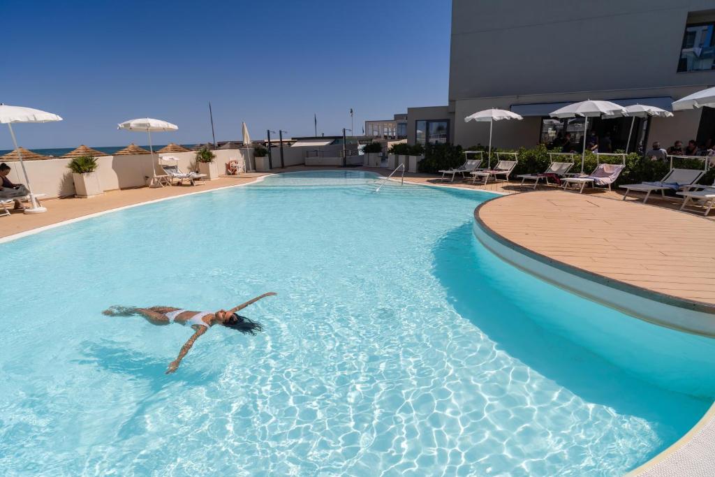 Peldēšanas baseins ar saulē pludojošu sievieti un sauļošanās krēsliem ap to, ideāla vieta atpūtai. Skats uz debesi un jūru fona rada relaksējošu atmosfēru.