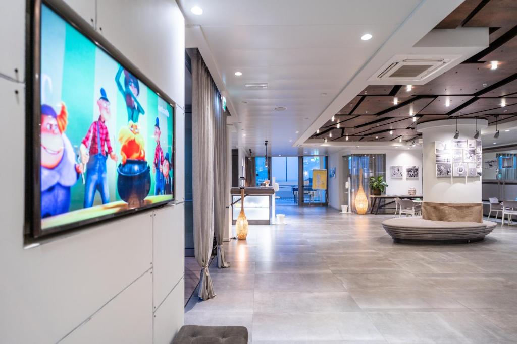 Mūsdienīga viesnīcas recepcija ar ērtu dīvānu un animācijas filmu attēlu uz ekrāna. Telpai ir gaiša un atklāta atmosfēra, ar stilīgiem apgaismojuma risinājumiem.