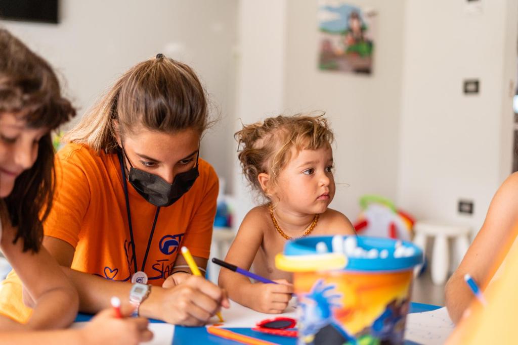 Bērni kopā ar pieaugušo radoši zīmē un krāso, izmantojot krāsainus zīmuļus un marķierus, kamēr viņi sēž pie galda. Telpa ir gaiša un draudzīga, radot radošu atmosfēru.