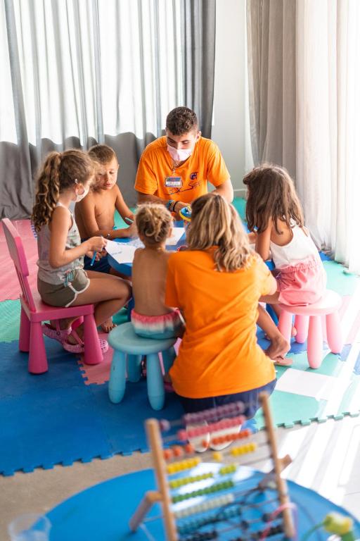 Bērni un pieaugušais radošā aktivitātē, zīmējot pie galda ar krāsainām krēsliem. Telpa ir gaiša un draudzīga, ar mīkstām paklāju segām.