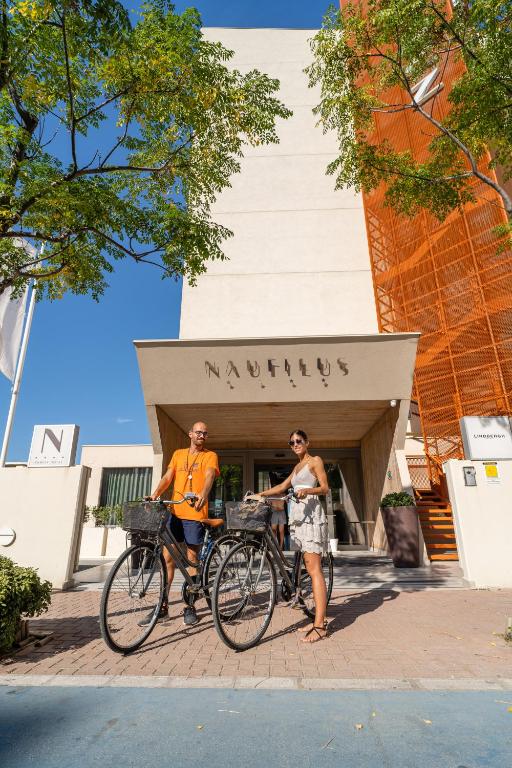 Divi cilvēki stāv pie viesnīcas "Nautilus", turēdami divus velosipēdus. Viesnīca ir moderna ar gaišām sienām un apzaļumotu teritoriju.