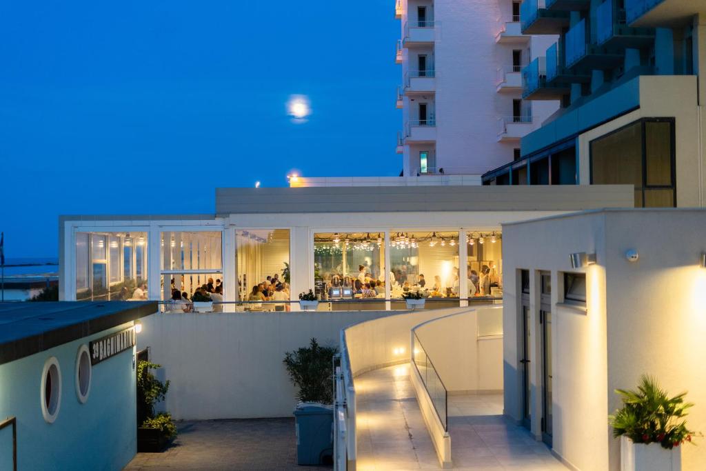 Moderns restorāns ar stiklotām sienām, kur iekšā redzama cilvēku grupa, kas baudī ēdienus. Fons ir nakts debesis ar mēnesi un tuvu esošu viesnīcu.