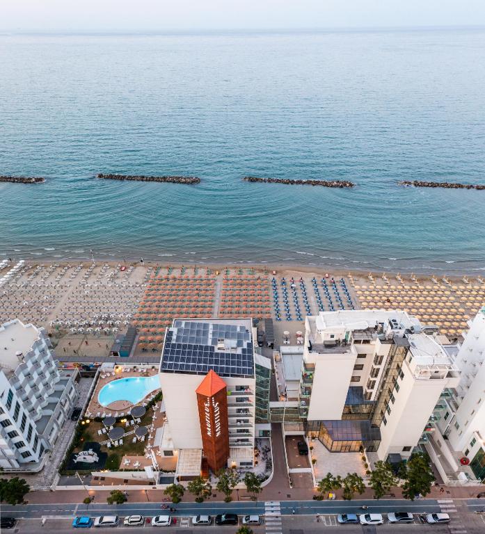 Kūrorts pie jūras ar peldbaseinu un saulessargu rindiņām pludmalē. Skats uz mierīgu jūru un piekrasti, kur redzamas viesnīcas un sauļošanās vietas.