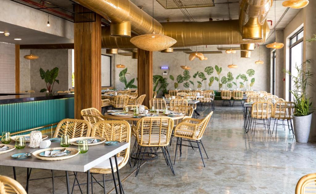 Mūsdienīgs restorāna interjers ar rattan krēsliem, elegantiem galdiem un augu motīviem uz sienām, radot patīkamu un viesmīlīgu atmosfēru.