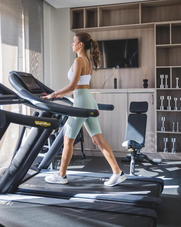 Sieviete vingro uz skrejceliņa modernā sporta zālē, apgaismotā telpā ar fitnesa aprīkojumu un televizoru fona.
