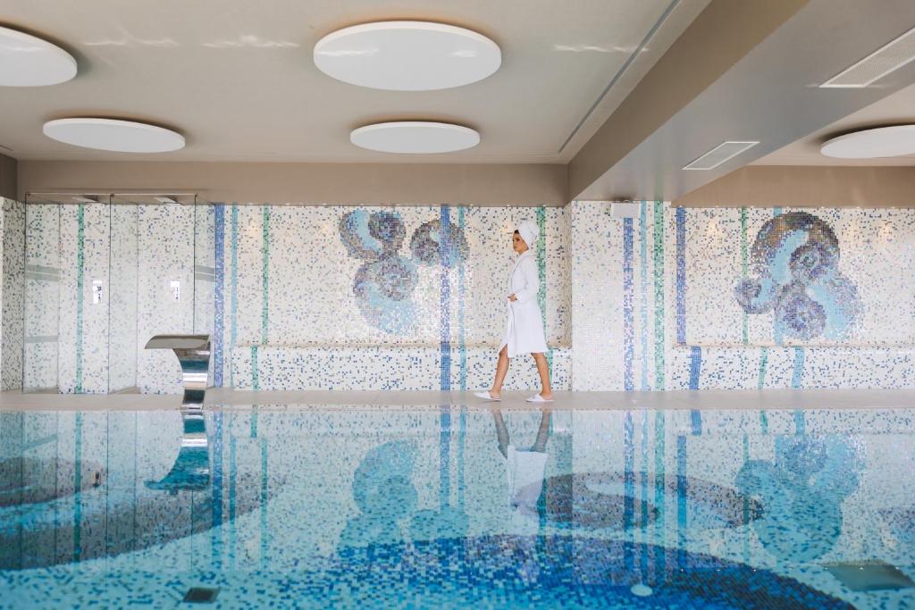 Elegants spa interjers ar mozaīkas sienām un mierīgu ūdens virsmu, kurā staigā apmeklētājs baltā halātā un dvielī. Ideāla vieta relaksācijai un atpūtai.