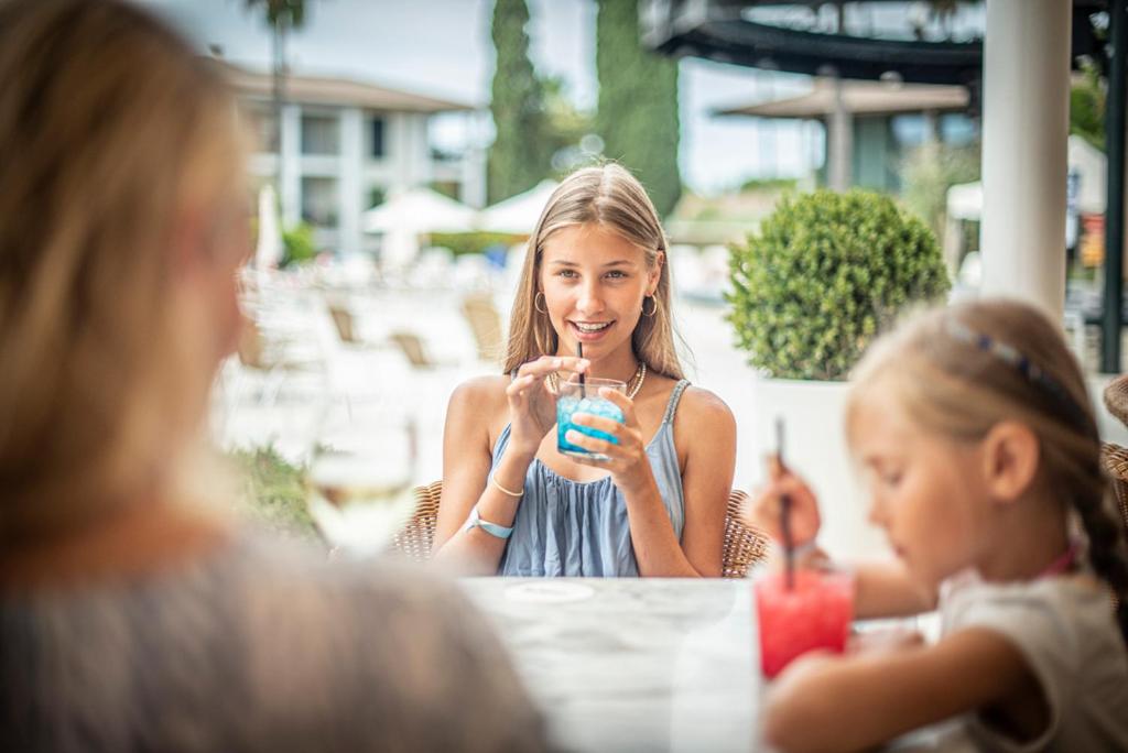 Jaunā sieviete sēž kafejnīcā, turot rokās dzērienu, kam ir spilgti zilā krāsa. Blakus viņai ir mazā meitene, kas ēd desertu. Fons ir vasarīgs, ar atpūtas zonu un zaļumiem.