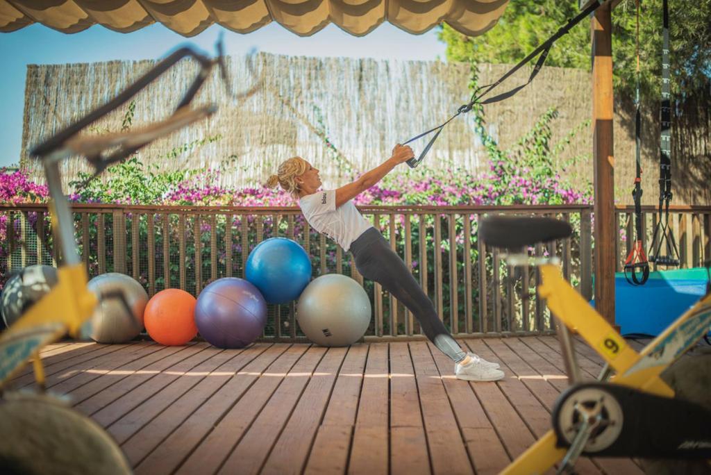 Sieviete vingro sporta zālē, izmantojot trenažierus un elastīgas lentes, fonā redzami dažādi vingrošanas bumbas.