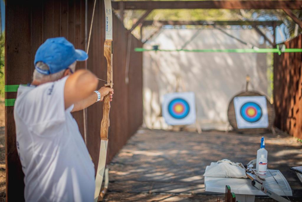 Arbaleta šaušanas treniņš, kurā dalībnieks koncentrējas uz mērķiem uz šautuves. Fons ir dabisks, ar diviem krāsainiem mērķiem un šaušanas aprīkojumu uz galda.