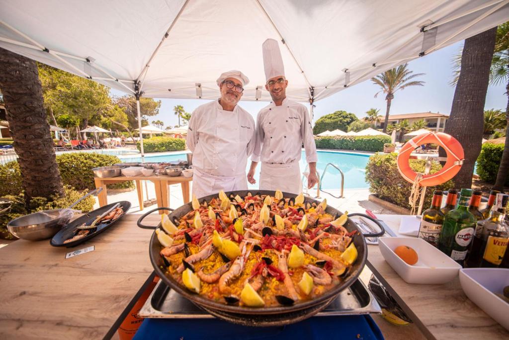 Šefpavāri stāv pie lielas paeljas pannas, kas piepildīta ar jūras veltēm un citroniem, fona ir peldbaseins un palmām.