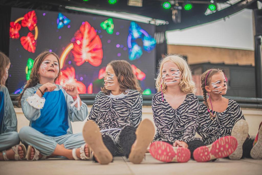 Bērni, tērpti zebra raksta kostīmos ar sejas zīmējumiem, sēž uz skatuves, kamēr fonā ir krāsains gaismas ekrāns.