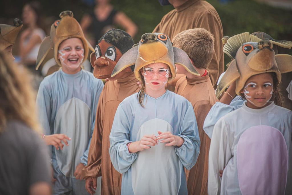 Bērni tērpušies dzīvnieku kostīmos, stāv grupā un gaida savu kārtu uz skatuves. Smieklīgas cepures un krāsains grims piešķir jautru atmosfēru pasākumam.