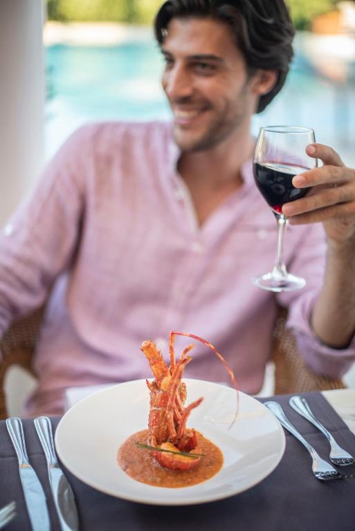 Vīrietis, kas sēž pie galda, tur glāzi sarkanvīna un smaida, kamēr priekšā viņam ir elegantā pasniegšanā izlikts ēdiens ar garnelēm. Fons ir relaksējoša atmosfēra ar baseinu.