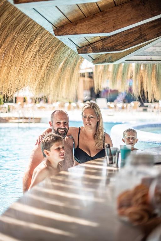 Ģimene izbauda laiku pie baseina, sēžot pie bāra zem salmu jumta, ar bērniem, kas priecīgi skatās uz kameru.