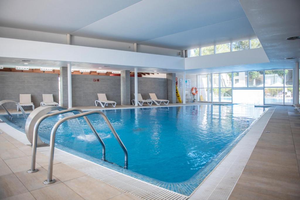 Iekštelpu peldbaseins ar skaidru ūdeni un atpūtas krēsliem, apgaismots ar dabīgu gaismu no logiem.