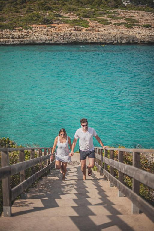 Pāris kāpj augšup pa kāpnēm, kas ved uz pludmali ar skaidru, zilu ūdeni un zaļām krasta augiem fonā.