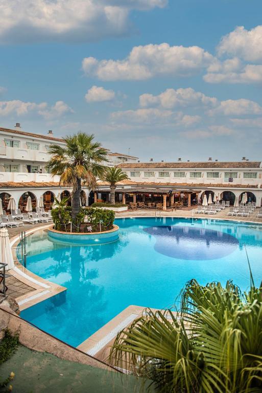 Viesnīcas baseins ar palmām un sauļošanās krēsliem, apkārtne ar ēkām un zilu debesu fonu. Ideāla vieta atpūtai un relaksācijai.