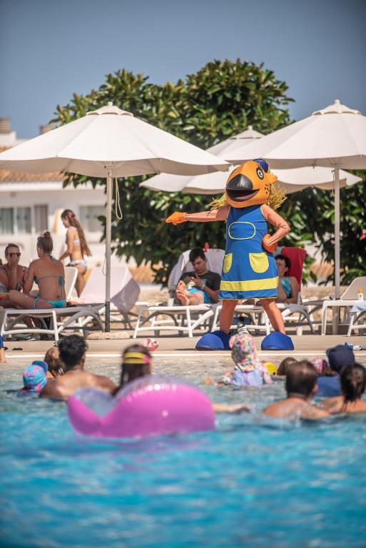 Baseina izklaides pasākums ar personāžu, kas tērpies zivju kostīmā, un cilvēkiem, kas atpūšas pie baseina. Rozā piepūšamā vaļa redzama ūdenī.