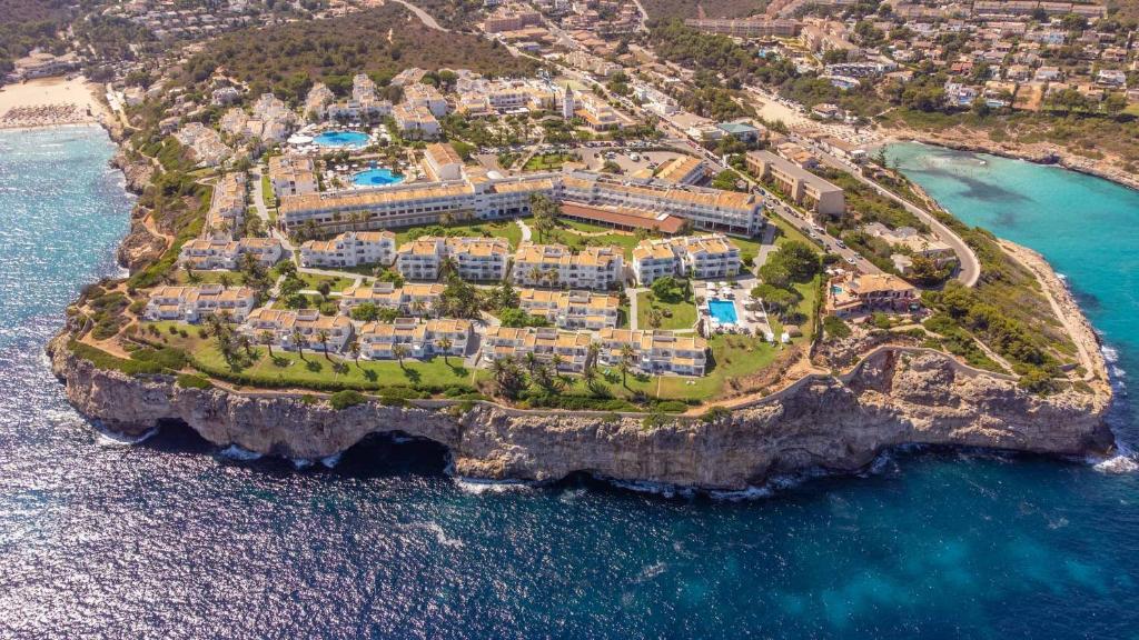 Aviācijas skats uz kūrortu, kas izvietots uz klints malas pie jūras, ar zaļiem laukumiem un baseiniem. Apkārt redzami pludmales un apkārtējā ainava.