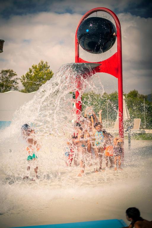 Ūdens atrakciju parks ar bērniem, kas priecīgi spēlējas zem milzīgas ūdens kritiena, kas iznāk no sarkanas struktūras.