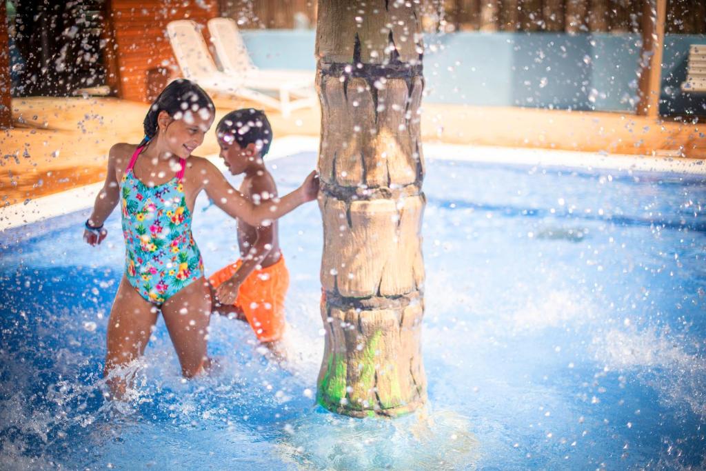 Bērni izbauda ūdens priekus peldbaseinā, spēlējoties ap koka stabu, ko aplido ūdens strūklas.