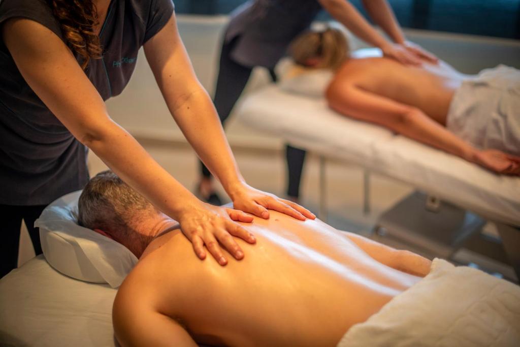 Masāžas sesija relaksējošā vidē, kur profesionāla masiere sniedz pakalpojumu, kamēr divi klienti guļ uz masāžas galdiem.