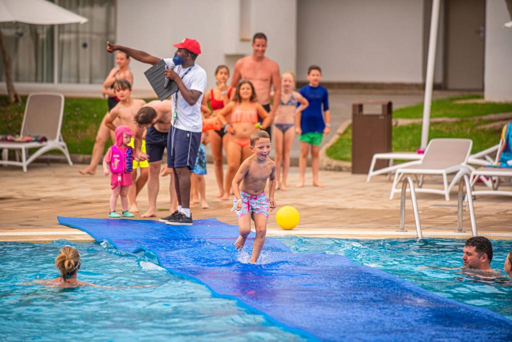 Bērns skrien pa ūdens slīdošo plati baseinā, priecīgi turēdams bumbu, kamēr citi bērni un pieaugušie vēro no malas. Tiek organizēta jautra aktivitāte pie baseina.