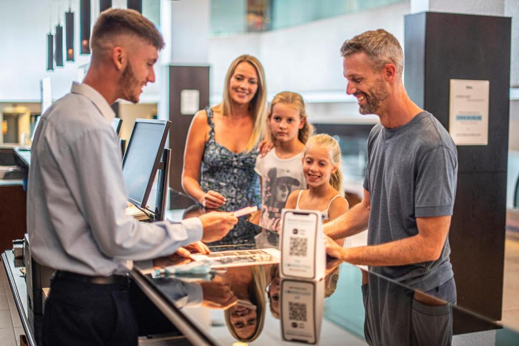 Ģimenes ierašanās viesnīcas reģistratūrā, kur viesmīlis apkalpo viesus, kamēr bērni skatās ar interesi.