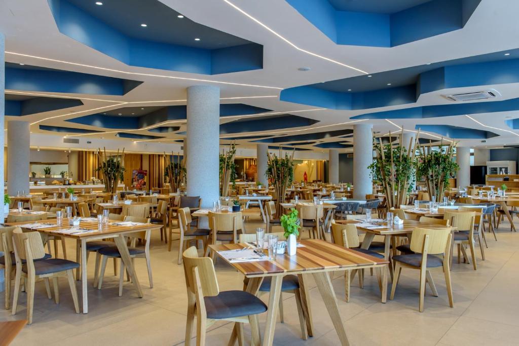 Mūsdienīgs restorāns ar elegantu interjeru, kurā ir koka galdi un krēsli, kā arī zaļi augi, kas rada patīkamu atmosfēru. Gaismas un krāsu kombinācijas piešķir telpai dinamiku un svaigumu.