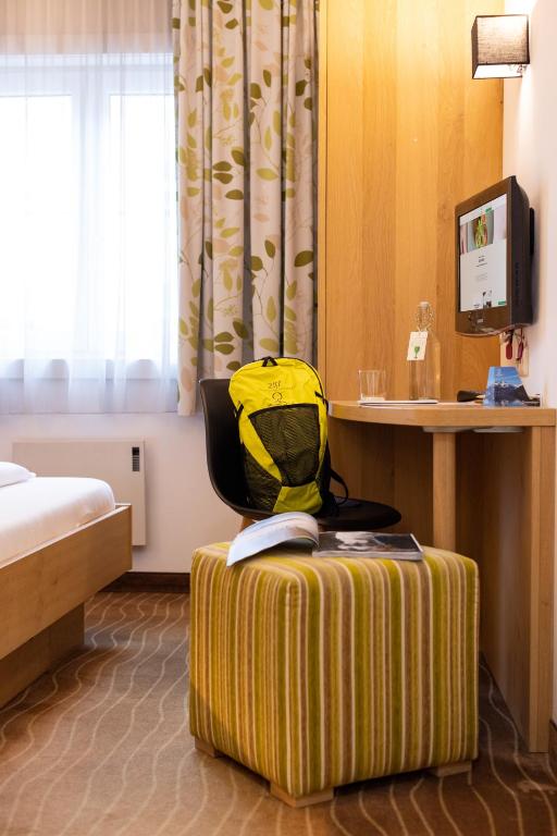 Mūsdienīgs viesnīcas numurs ar koka mēbelēm, darba galdu un krēslu, dzeltenu mugursomu un televizoru. Logā ieplūst dabiska gaisma, radot patīkamu atmosfēru.