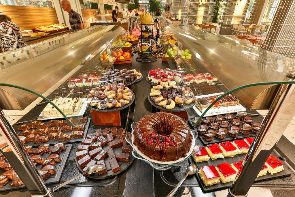 Deserts galdu bufete ar dažādām kūkām, saldumiem un augļiem, izkārtoti uz melna galda. Aiz galda redzami apmeklētāji un stilīga restorāna atmosfēra.