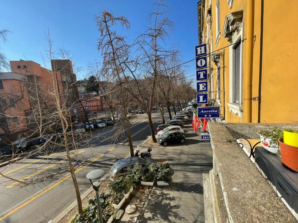 Skats no viesnīcas loga uz ielu, kurā redzami stāvoši automobiļi un koka zari, kas zaudējuši lapas. Uz viesnīcas izkārtnes redzams nosaukums "Aurelia".