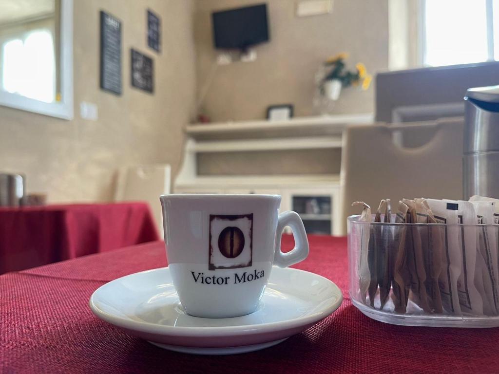 Kafijas krūze ar "Victor Moka" logo uz balta apakštase, novietota uz sarkanas galdautas. Fonā redzama kafejnīca ar dekorācijām un ērtām mēbelēm.