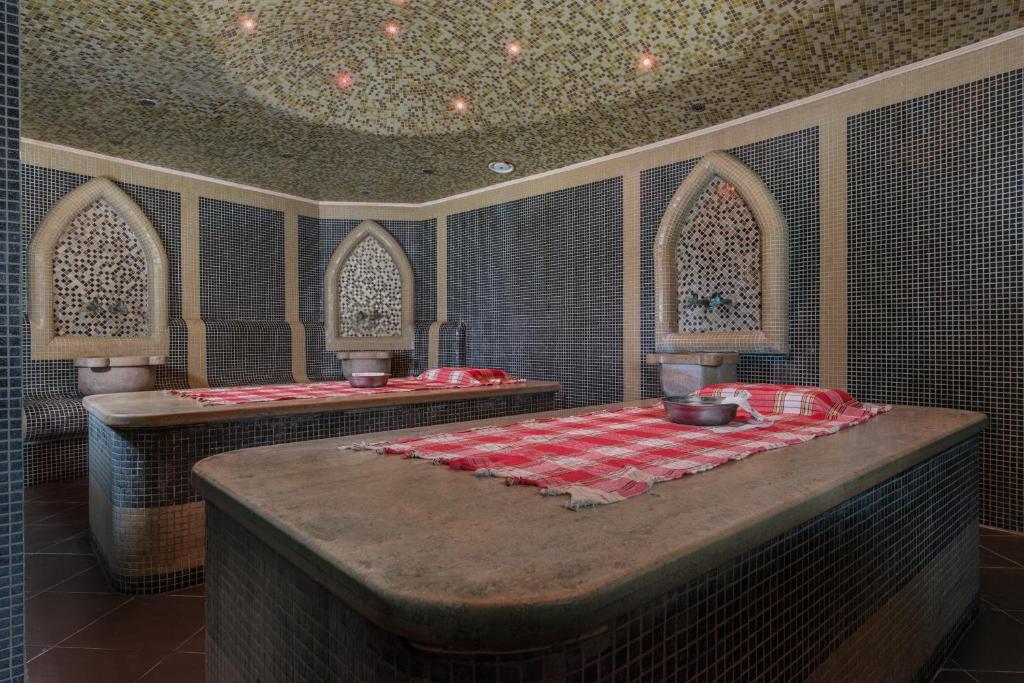 Spa telpa ar mozaīkas sienām un dekoratīvām nišām, kur atpūtas galdi ir klāti ar sarkanu un baltu pārklāju. Telpā ir relaksējoša atmosfēra.